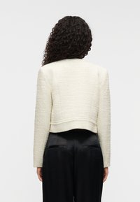 Λευκό υφασμάτινο κοντό σακάκι με μακριά μανίκια, με ίσια γραμμή στο τελείωμα και απαλά ραφικά σχέδια. Φορεμένο πάνω από μαύρο παντελόνι.