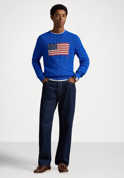 Blue cable-knit sweater featuring a prominent American flag motif on the front, paired with dark denim trousers and brown boat shoes.