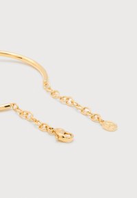 Βραχιόλι με χρυσή αλυσίδα, κούμπωμα με κροκόδειλο και μικρή εξαγωνική ετικέτα, τοποθετημένο σε απλό λευκό φόντο.