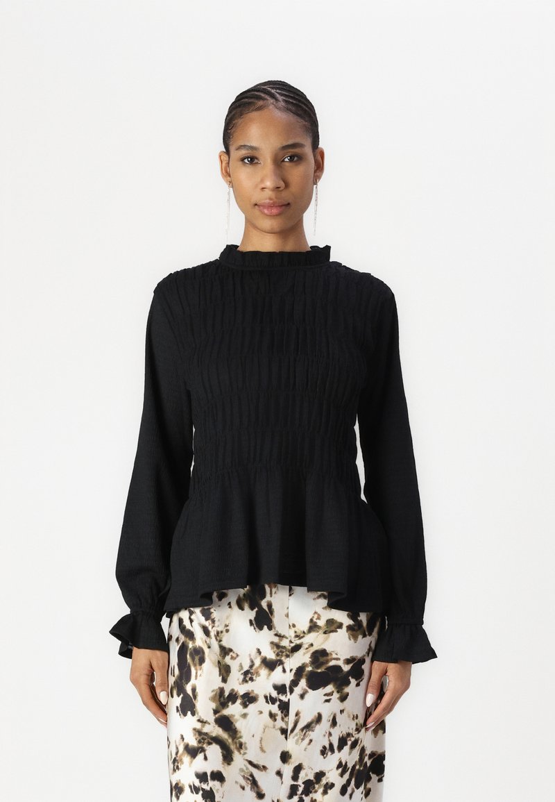 Black long-sleeve top with a smocked texture and ruffled neckline. Features pleated bodice and peplum hem. Worn with a patterned beige skirt.