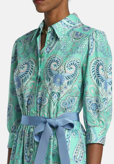 Woman wearing a turquoise paisley floral dress with button front, puffed sleeves, and a tied blue waist belt.