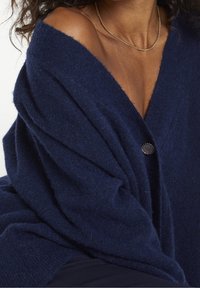 Marinblå oversized cardigan i mjukt stickat tyg, med en djup V-ringning, draperad design och en rund knapp fram.