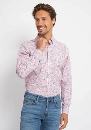 Man smiling and touching his chin, wearing a pink patterned long-sleeve shirt and blue jeans against a plain white background.