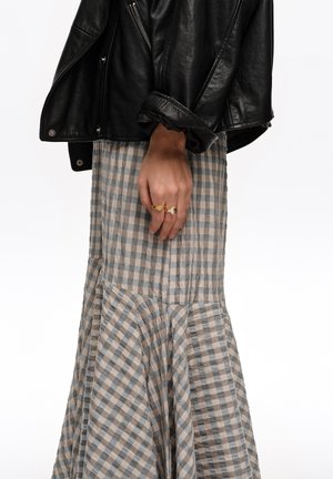 Mano apoyada al costado con anillos, chaqueta de cuero negra con puños doblados y falda a cuadros azul y beige de cintura alta.