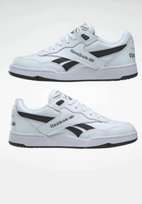 White leather sneakers with black accents, featuring a round toe, lace-up front, and Reebok logos on the side and heel. Textured rubber sole.