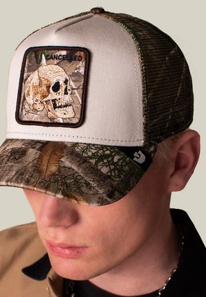 Hombre con gorra beige y de camuflaje con parche de calavera y texto "CANCELLED" en el frente, chaqueta beige, mirando hacia abajo.