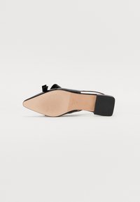Zapato de tacón alto negro con punta afilada, hecho de cuero suave. Presenta un acento de lazo y un tacón cuadrado con una suela texturizada.