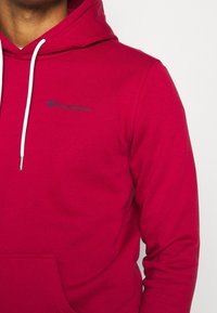 Felpa rossa realizzata in tessuto morbido, con tasca anteriore, lacci bianchi e un piccolo logo blu di Champion sul petto.