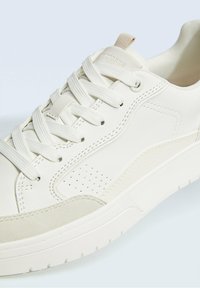 Sneaker bassa bianca con suola testurizzata, lacci bianchi e piccole aperture di ventilazione sul pannello laterale.