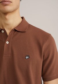 Brūns polo krekls ar teksturētu audumu, diviem pogām pie apkakles un mazu melnbaltu izšūtu putna logotipu uz krūtīm.