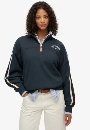 Sweat-shirt bleu marine avec une demi-fermeture éclair et un logo brodé, présentant des rayures crème et bourgogne sur les manches. Superposé à une chemise rayée.