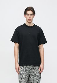 Młody mężczyzna o średniej długości ciemnych włosach nosi czarną koszulkę Nike i białe spodnie w wzory na jasnym, jednolitym tle.