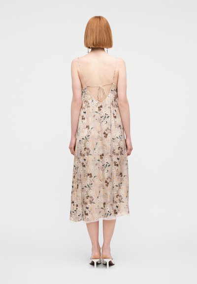 Robe nuisette florale en tissu doux et transparent. Comprend des bretelles fines, un détail noué au dos, et une couleur beige clair avec des motifs botaniques.