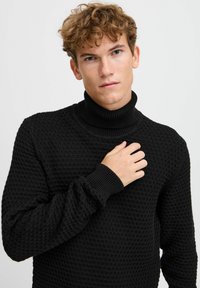 Suéter negro de cuello alto con textura y puños acanalados. Presenta un patrón de punto en forma de panal y un diseño ajustado que proporciona calidez y comodidad.