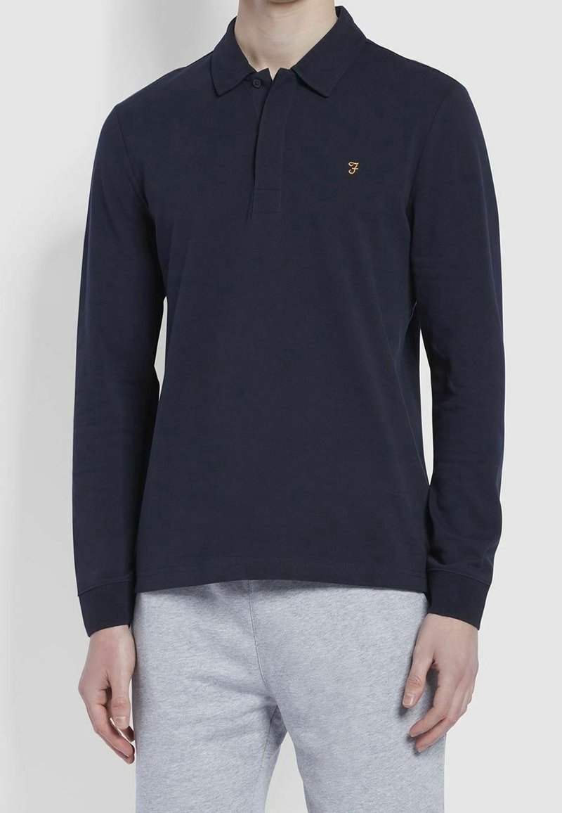 Námořnická modrá polokošile s dlouhým rukávem, tříknoflíkovou légou a vyšitým logem zlaté barvy na levé straně hrudi. Hladká látka, klasický střih.