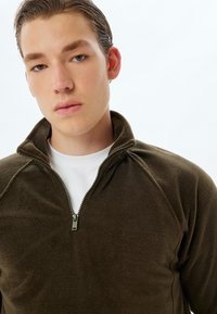 Sudadera de forro polar marrón con cuello alto y media cremallera. La tela tiene una textura suave. Combinada con una camiseta de cuello redondo blanca debajo.
