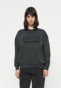 Donna con capelli scuri alla lunghezza delle spalle, che indossa un felpa sportiva Rip Curl Surf grigio scuro e pantaloni abbinati scuri, in piedi contro uno sfondo semplice.