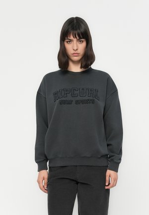 Donna con capelli scuri alla lunghezza delle spalle, che indossa un felpa sportiva Rip Curl Surf grigio scuro e pantaloni abbinati scuri, in piedi contro uno sfondo semplice.