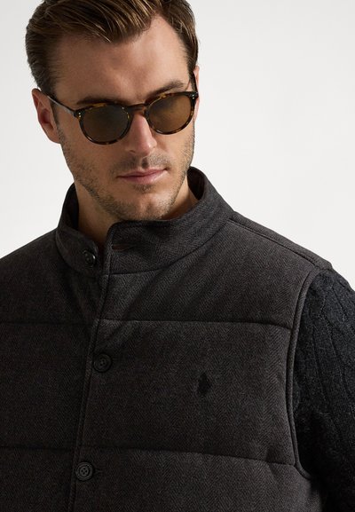 Vestă gri cu model în formă de herringbone, cu guler înalt, închidere cu nasturi și mâneci negre texturate în contrast. Ochelari rotunzi, întunecați, completează ținuta.