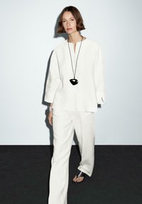 Linnedragt i off-white, med en knap-op top og brede bukser. Accepteret med en sort pendant halskæde og en kraftig sølvur.