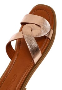 Sandalias de tiras metálicas en oro rosa con un diseño trenzado, plantilla de espuma marrón y suela flexible. Textura elegante y suave.