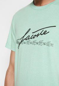T-shirt vert menthe à manches courtes avec un logo imprimé noir et plusieurs graphiques stylisés de crocodiles sous le logo.