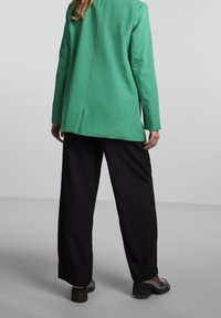 Femme portant un blazer vert vif, un pantalon noir ample et des chaussures à talons noirs, se tenant devant un fond gris uni.