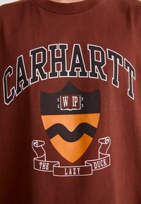 Czerwono-brązowa bawełniana koszulka z nadrukiem "CARHARTT" na białym tle, nad wzorem tarczy z napisem "WIP", pomarańczowymi akcentami i wstęgą z napisem "THE LAZY DUCK".