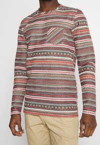 Camicia a maniche lunghe con un motivo etnico multicolore, caratterizzata da design geometrici in rosso, beige e nero, e una tasca sul petto. Materiale in cotone.