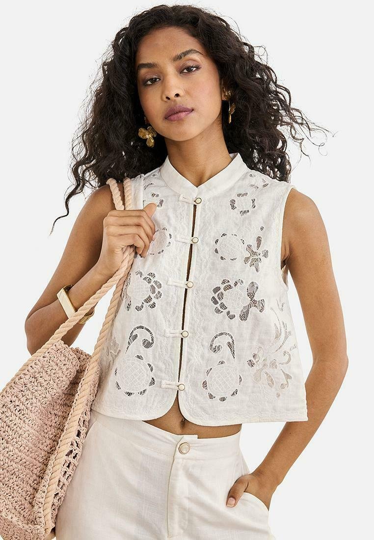 Mujer con top sin mangas blanco bordado con botones y pantalones claros, llevando un bolso tote beige tejido sobre el hombro.
