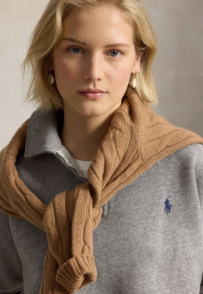 Blond meisje draagt een grijze trui met rits en blauw logo en een beige kabelgebreide trui over haar schouders.
