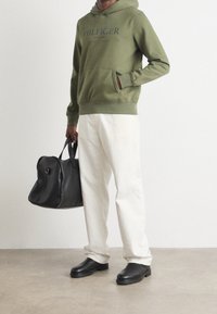Felpa verde oliva con tasca frontale e logo blu navy, abbinata a pantaloni color crema. Una borsa a mano nera in pelle e scarpe nere completano l'outfit.