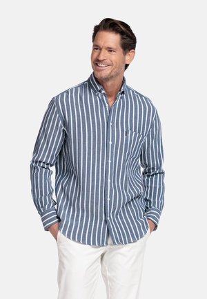 Lachende man die een blauw-wit gestreept overhemd met knopen en kraag draagt, met witte broek, handen in zijn zakken, tegen een effen achtergrond.