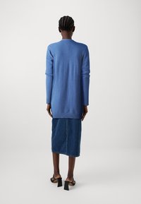 Cárdigan azul de manga larga hecho de material suave, con un dobladillo acanalado, combinado con una falda de mezclilla y sandalias negras de tacón cuadrado.