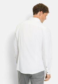 Mann, der ein langärmliges weißes Hemd und graue Hosen trägt, von hinten gezeigt, stehend vor einem schlichten hellen Hintergrund.