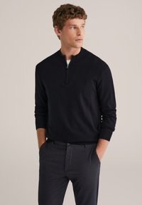 Pull-over noir à col demi-zip, poignets et ourlet côtelés, porté sur une chemise blanche, assorti à un pantalon gris foncé.