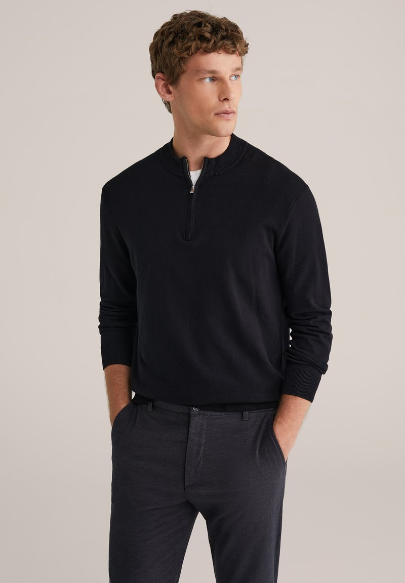 Pull-over noir à col demi-zip, poignets et ourlet côtelés, porté sur une chemise blanche, assorti à un pantalon gris foncé.