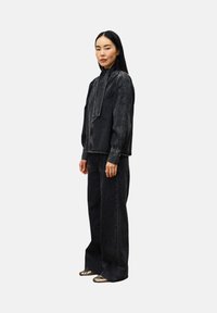 Frau steht in einer schwarzen Denim-Bluse mit langen Puffärmeln, schwarzen weitgeschnittenen Jeans und metallisch glänzenden spitz zulaufenden Schuhen.