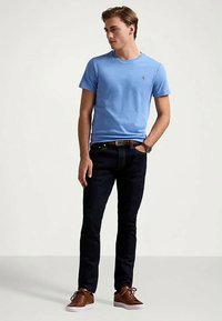 Νεαρός άνδρας που στέκεται φορώντας ανοιχτό μπλε κοντομάνικο πουκάμισο, σκούρες τζιν παντελόνες, καφέ δερμάτινα παπούτσια και ρολόι, κοιτώντας προς τα αριστερά.