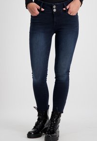 Eng passgenaue Jeans aus dunklem Denim mit mittelhohem Bund, zwei vorderen Taschen und doppeltem Knopfverschluss, kombiniert mit schwarzen Schnürstiefeln.