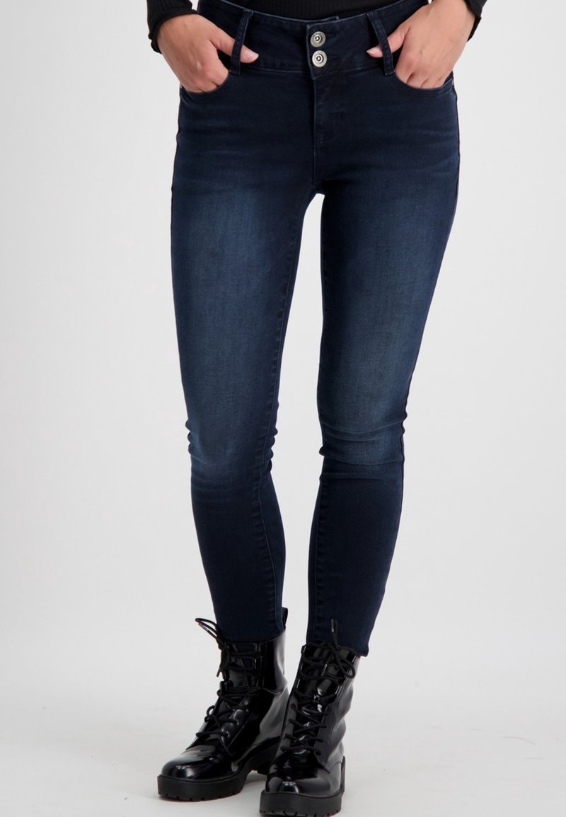 Eng passgenaue Jeans aus dunklem Denim mit mittelhohem Bund, zwei vorderen Taschen und doppeltem Knopfverschluss, kombiniert mit schwarzen Schnürstiefeln.
