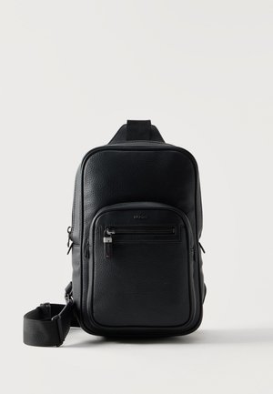 Sac bandoulière en cuir noir texturé avec poche zippée à l'avant, sangle réglable et logo de marque discret sur fond blanc.