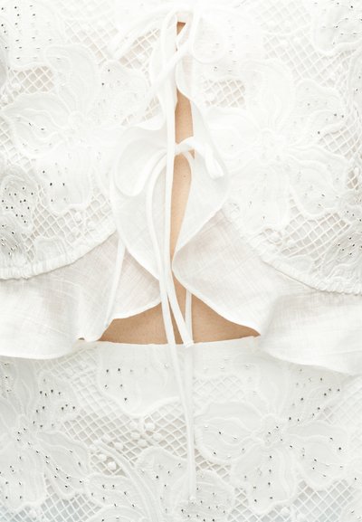 Λευκό δαντελωτό ένδυμα με λουλουδένια μοτίβα και μικρές ασημένιες χάντρες, με λεπτομέρεια δεμένης βολάν μπροστά πάνω στο δέρμα.