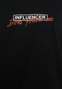 Crna pamučna majica s bijelim blokovnim natpisom "INFLUENCER" i crvenim kurzivnim natpisom "Ne slijedi me" za vizualni kontrast.