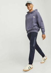 Sudadera morada con bolsillo canguro y logo, pantalones de chándal azul marino con texto de la marca, combinados con zapatillas blancas con acentos verdes y rojos.