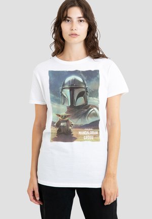 Woman wearing white T-shirt featuring the Mandalorian helmet and Grogu standing on desert terrain under a cloudy sky.