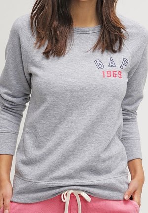 Vrouw draagt een lichtgrijze sweater met lange mouwen en 'GAP 1969' op de borst, gecombineerd met roze joggingbroek met een witte trekkoord.