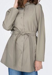 Femme portant un imperméable beige ceinturé avec des boutons-pression sur un jean bleu, main touchant les cheveux près du visage.
