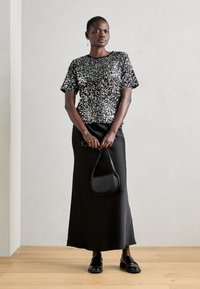 Haut en sequins noirs à manches courtes associé à une jupe longue noire. Le mannequin tient un petit sac à main noir incurvé et porte des mocassins noirs.