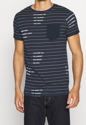 Man wearing navy blue striped short-sleeve t-shirt with white crossed-out text and a chest pocket, paired with dark jeans.